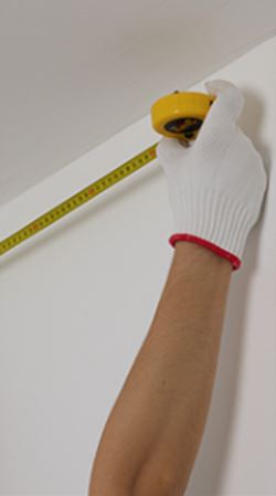 Gloved hand measuring wall height with yellow tape measure near ceiling